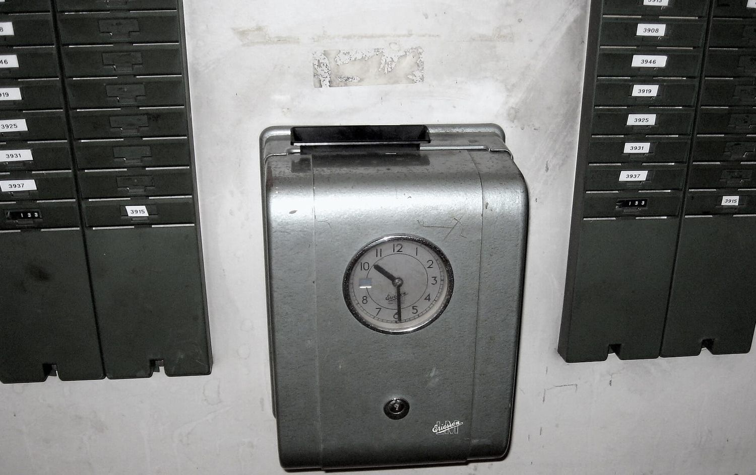 Alte mechanische Stempeluhr an einer Wand zwischen zwei Regalen mit nummerierten Stempelkarten