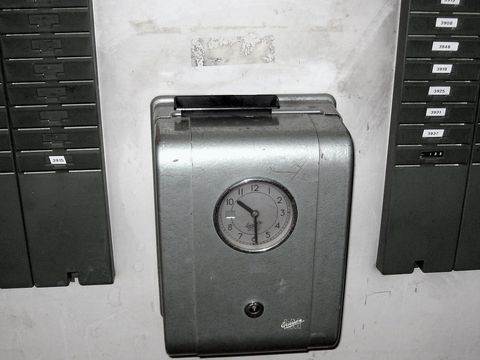 Alte mechanische Stempeluhr an einer Wand zwischen zwei Regalen mit nummerierten Stempelkarten